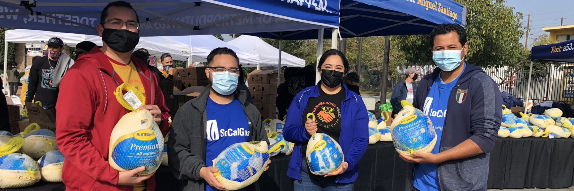 SoCalGas volunteers holding frozen turkeys with tables of frozen turkeys behind them